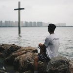 man in white shirt sitting on brown rock near body of water during daytime