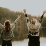 two women hands up standing beside body of water