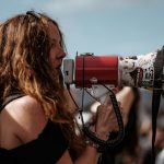 selective focus photography of woman wearing black cold-shoulder shirt using megaphone during daytime